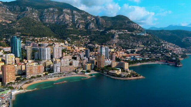 Aerial drone view of the Larvotto beach district in Monaco with residential towers, sandy bays, breakwaters, seafront promenade, the Mediterranean coastline and mountains toward Roquebrune.