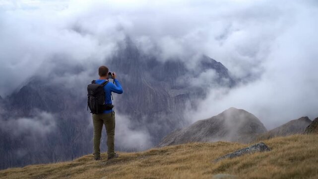 Person with backpack aims a handheld camera at cloud-covered mountain peaks from an alpine ridge, standing above muted meadow terrain as mist drifts between rocky slopes