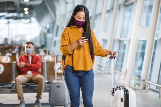A woman in a face covering uses her smartphone and holds a passport while standing with luggage in a modern airport departure lounge