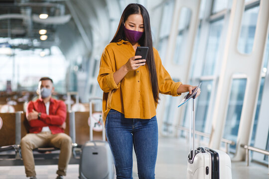 A young woman wearing a protective mask checks her phone while holding a passport and suitcase in a modern airport terminal