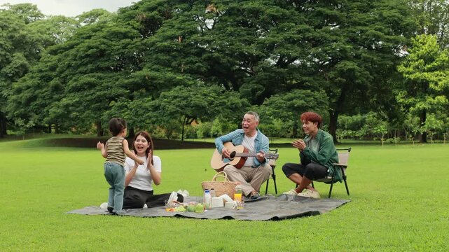 GrandFather playing acoustic guitar for child during picnic. Asian family enjoy outdoor picnic with food and drinks in park sunny day, outdoor activitie with daughter father, mother grandfather