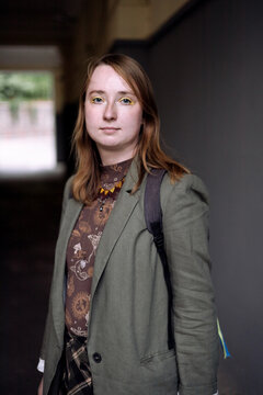 a serious but confident young businesswoman standing with crossed arms against a plain dark grey background. She is wearing a professional olive-green blazer over a pattern blouse, looking directly at