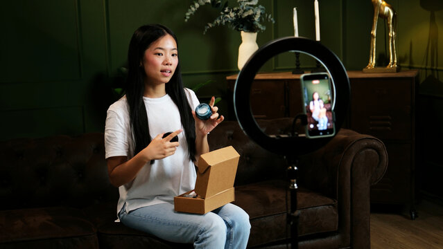 Young Asian woman unboxing cosmetic products while streaming live video at home