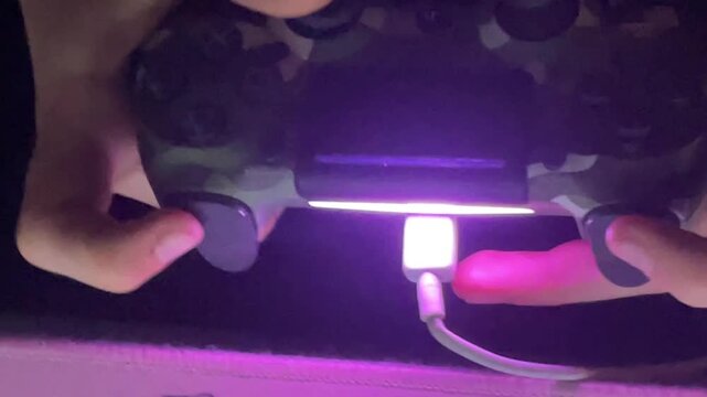 Gaming closeup hands with controller. Vertical charging controller with glowing purple led, usb connector plugged into port, steady grip and button pressure