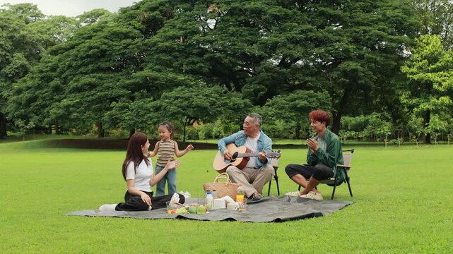 GrandFather playing acoustic guitar for child during picnic. Asian family enjoy outdoor picnic with food and drinks in park sunny day, outdoor activitie with daughter father, mother grandfather