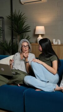 Mature woman learning computer skills from her asian adopted granddaughter on a couch