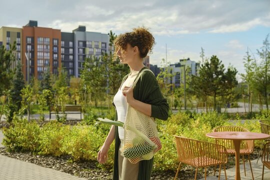 Woman walks through park while holding reusable mesh bag filled with vegetables. Green trees and buildings are in the background