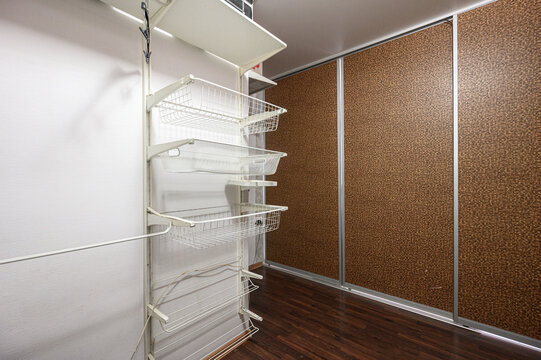 Compact storage area features white metal wire shelves, sliding brown patterned wardrobe doors, dark wooden floor, minimalist design, efficient space utilization