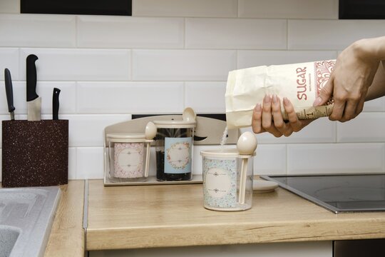 A person is pouring sugar into a container in a kitchen. The countertop is made of wood and has jars for sugar and other items. Several kitchen tools are visible in the background