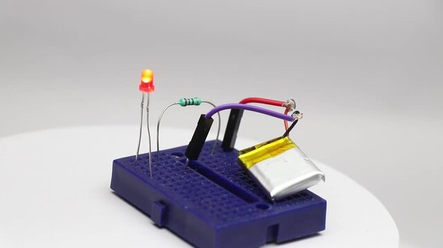 Breadboard with small red LED glowing connected to lithium polymer battery power through a resistor rotating to show the details of the circuit