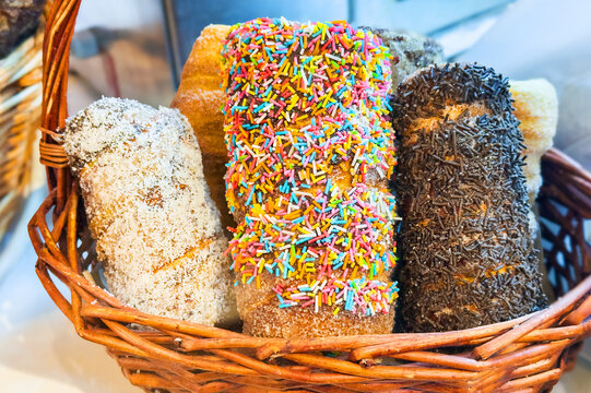 Assorted bakery pastries coated in colorful sprinkles, coconut, and chocolate vermicelli displayed together in a rustic wicker basket at a market.