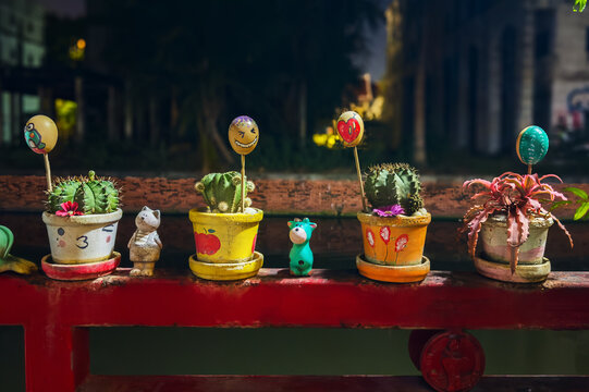 Whimsical hand painted face eegs on sticks decorate small potted cacti and succulents on red railing at night in garden cafe. Colorful painted egg shells characters with funny expressions.