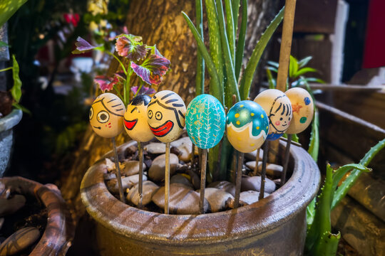 Row of hand painted egg shells with faces on sticks standing among aloe and coleus plants in a rustic clay pot, cheerful garden decoration. Colorful painted empty eggs with cartoon expressions.