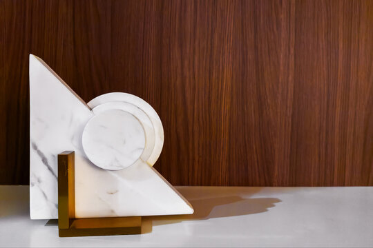 Abstract white marble sculpture with brass base standing against a warm wood paneled wall, minimalist modern home decor object. Elegant contemporary interior decoration. Copy space background.