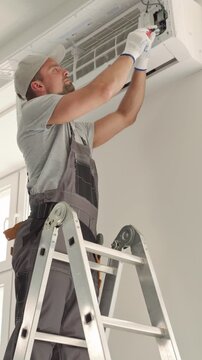 Technician performs HVAC repair on air conditioner. Worker on ladder services wall mounted unit indoors for cooling maintenance and safety. Essential climate control service for homes.