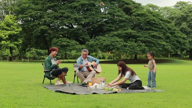 GrandFather playing acoustic guitar for child during picnic. Asian family enjoy outdoor picnic with food and drinks in park sunny day, outdoor activitie with daughter father, mother grandfather