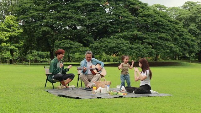 GrandFather playing acoustic guitar for child during picnic. Asian family enjoy outdoor picnic with food and drinks in park sunny day, outdoor activitie with daughter father, mother grandfather