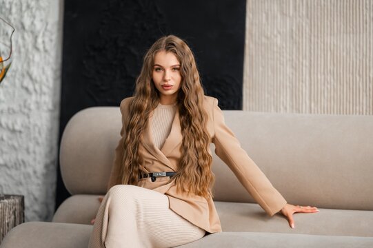 Smiling young woman with long wavy hair seated on modern neutral sofa wearing beige blazer and ribbed knit dress