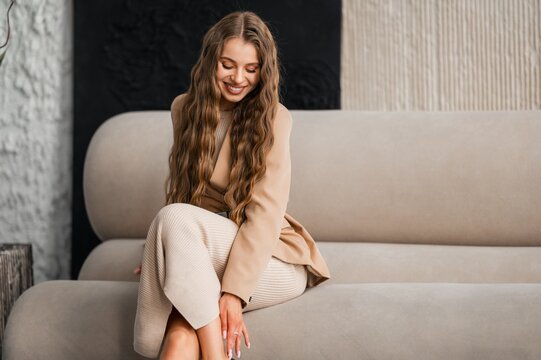 Portrait of woman in beige blazer and matching knit dress seated on light gray sofa in modern two tone interior with textured beige and black wall soft natural light