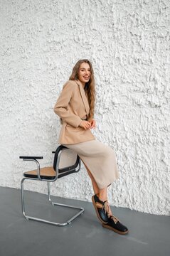 Smiling young woman in beige blazer and ribbed knit skirt leaning on metal chair, long wavy hair against textured white plaster wall in bright studio light