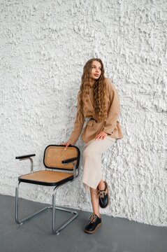 Young woman with long wavy light brown hair leaning on woven chair against textured white plaster wall wearing beige blazer and ribbed knit skirt in studio fashion portrait