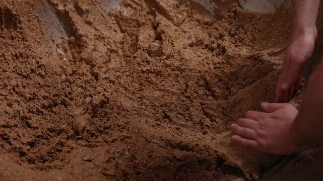 Hands work dense, dark rye bread dough inside a large wooden vessel, showing the rustic texture and traditional process of bread making