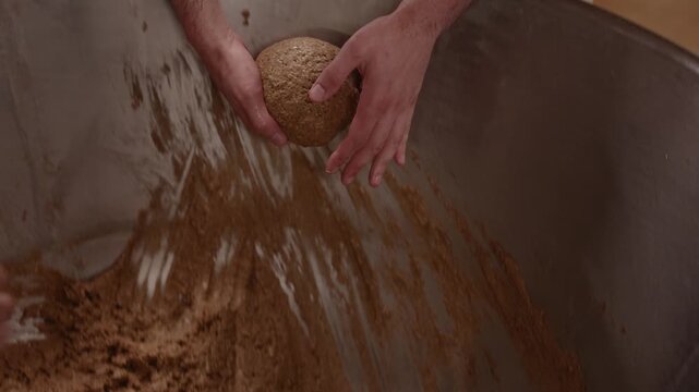 Hands work dense, dark rye bread dough inside a large wooden vessel, showing the rustic texture and traditional process of bread making