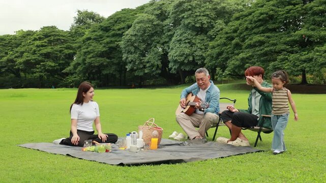 GrandFather playing acoustic guitar for child during picnic. Asian family enjoy outdoor picnic with food and drinks in park sunny day, outdoor activitie with daughter father, mother grandfather