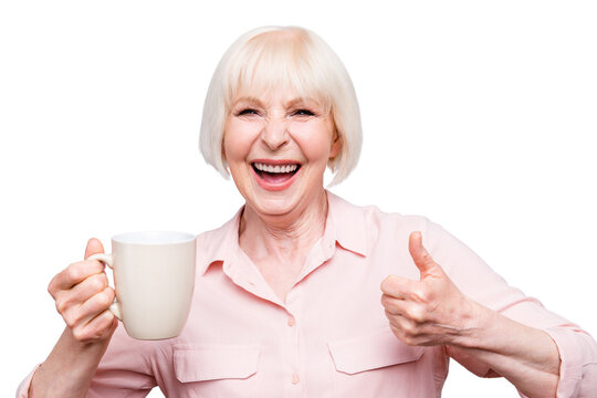 Portrait of old excited lady showing thumbs up gesture, smiling laughing, holding cup drinking coffee, tea, beverage on grey background, isolated