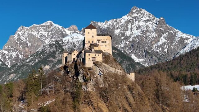 stone keep tower tarasp castle rises sheer rock pinnacle round turret linked rampart wall alpine peaks snow behind golden larch forest patches snow ground lower engadin switzerland aerial footage