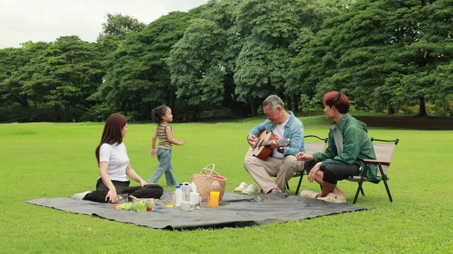 GrandFather playing acoustic guitar for child during picnic. Asian family enjoy outdoor picnic with food and drinks in park sunny day, outdoor activitie with daughter father, mother grandfather