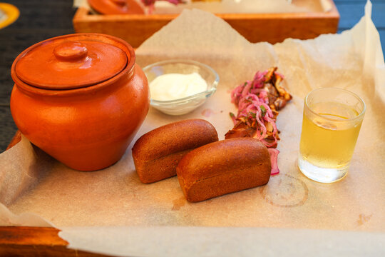 Hearty homemade soup served in a rustic clay pot, accompanied by small fresh bread rolls, crispy bacon pieces, and a cup of sour cream.