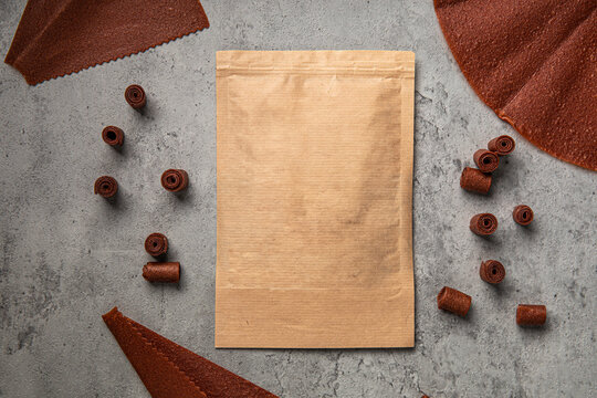 Natural fruit leather rolls and sheets arranged around craft paper ziplock pouch mock up on grey concrete background