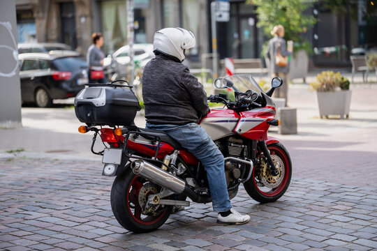 Helmeted rider sits on a motorcycle with a rear luggage case and one foot on cobblestones, illustrating personal motorized transport and urban road travel