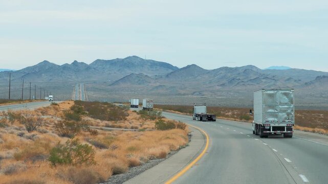 Several semi trucks driving in convoy on winding asphalt highway through arid desert landscape of american west, with distant mountains under cloudy sky. Transportation of goods and cargo