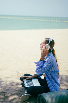 Woman relaxing with headphones while working on laptop at beach, blending remote work, music, and wellness lifestyle