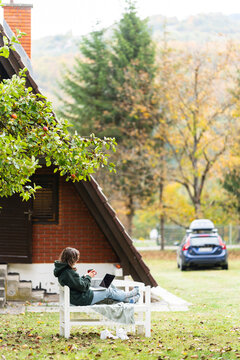 A person sitting outdoors on a wooden bench with a laptop during autumn in front of a rural house and a car.