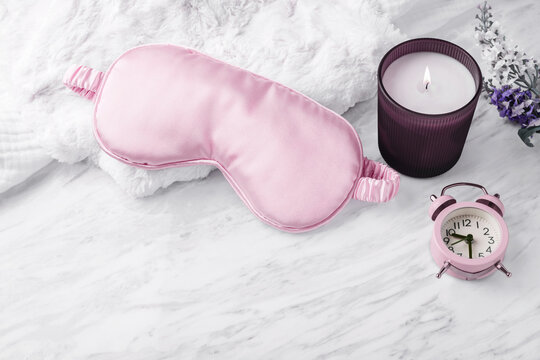 Pink eye mask with lit scented candle, alarm clock, and  flowers on marble surface
