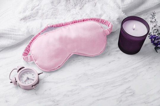 Silk sleep mask on soft fabric with aroma candle and alarm clock on marble background