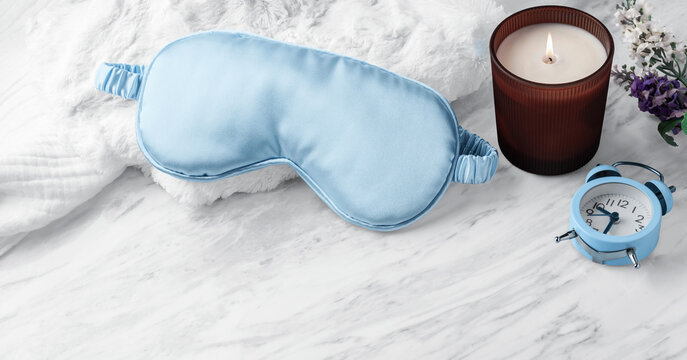 Light blue silk sleeping mask on soft fabric with aroma candle and alarm clock on marble background