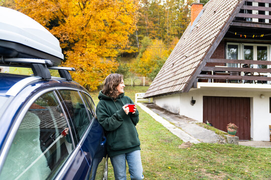 A woman leaning against a blue car with a red mug in her hand in front of a wooden A-frame house surrounded by colorful autumn forest