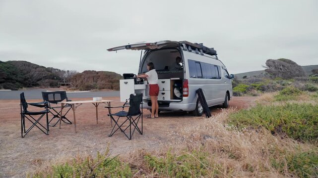Woman cooks by a van parked near shrubs on a cloudy day, with a small table and chairs set nearby, little girl is inside the van sitting on a bed