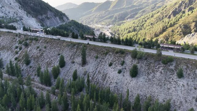 Cinematic top-down drone flyover directly above crest of massive embankment mudflow control dam with steep slopes, protective forest, and access road in Almaty, Kazakhstan at summer sunset