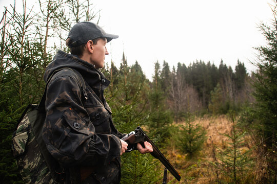 A hunter in a cap and camouflage jacket inspecting his shotgun while outdoors in an autumn forest edge