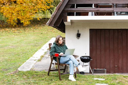 A woman sitting in a wooden chair outside an A-frame house with a laptop and a red mug during autumn