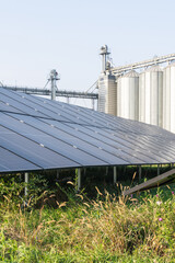 A ground-mounted solar panel array installed in a grassy field near an industrial agricultural facility with grain silos © scharfsinn86
