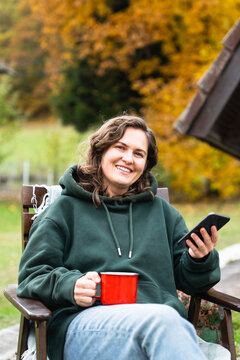A woman sitting in a wooden chair outside an A-frame house holding a smartphone and a red mug during autumn