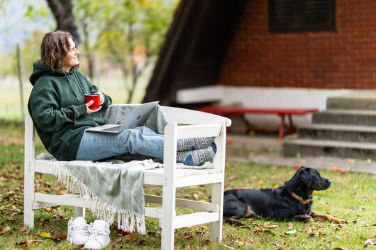 A woman sitting on a white wooden bench with a laptop and a red cup while a black dog lies nearby in a yard during autumn