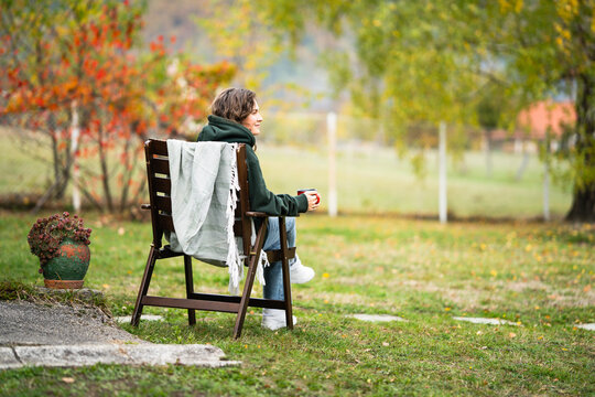 A woman sitting in a wooden chair with a blanket and a red mug, relaxing outdoors during autumn in a countryside garden