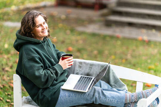 A woman sitting on a white wooden bench under an apple tree with a laptop and a red mug during autumn in a rural garden.
